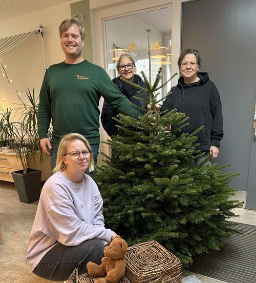 Herr Fichter hält einen Tannenbaum, den er gerade in den Kindergarten liefert. Eine Erzieherin, die Kitaleiterin und Frau Stiller von den Stadtwerken stehen neben dem Baum.